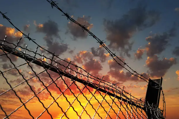 Barbed wire fence against a dramatic sky at sunset. The concept of restriction of freedom of movement, human rights violations.