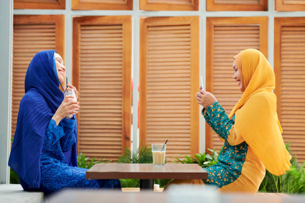 Cheerful young muslim woman in hijab taking photo of her friend with sweet milky cocktail on smartphone