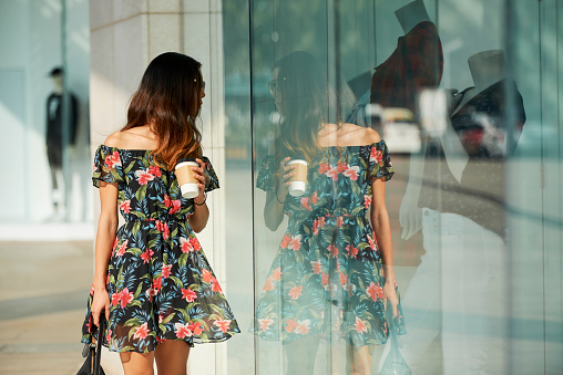 Attractive young woman walking in the street with take out coffee in her hands and looking at shop windows