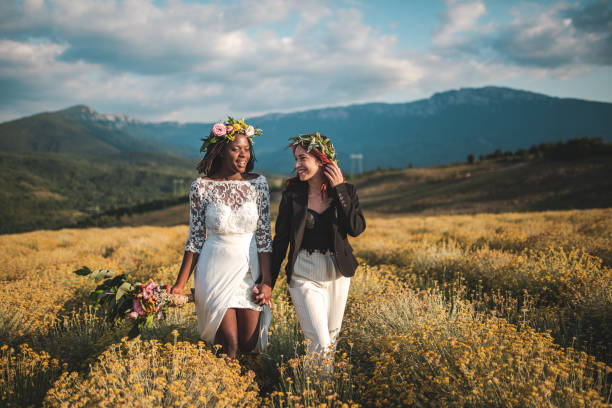 Cheerful married lesbians on their special day in nature Two beautiful, young women holding hands and walking on meadow on their wedding day eloping stock pictures, royalty-free photos & images