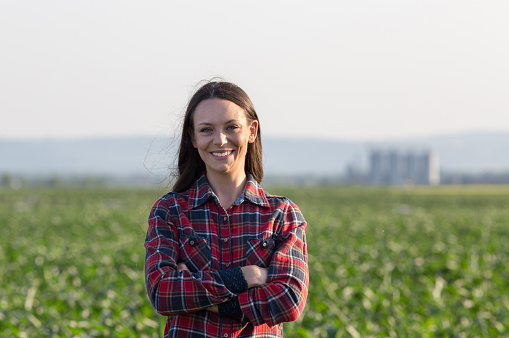 Portrait of pretty young farmer woman with crossed arms in corn field with grain silos in background