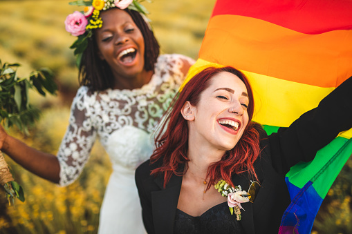 Married lesbian couple holding rainbow flag and celebrating their love happily, laughing and embracing