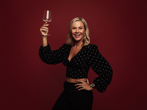 Woman cheering with glass of wine in studio against red\nPortrait of mid adult modern woman in studio