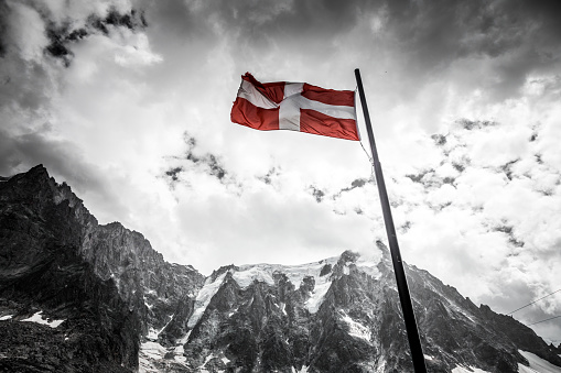 Desaturated color image depicting the red and white of the Danish national flag blowing in the wind in the middle of a rugged mountain landscape in the wilderness with a dramatic cloudscape. Room for copy space.