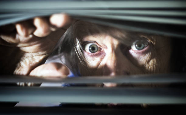 istock Frightened senior woman looking, wide eyed, through blinds 1168895721
