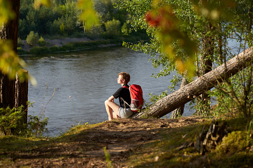 Caucasian short hairstyle woman with red backpack sitting on a log above river and looking faraway. Relaxation.