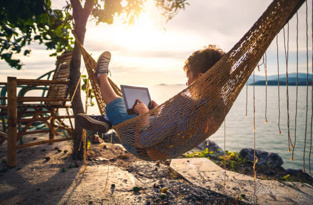 A boy uses digital tablet lying in hammock near sea at sunset