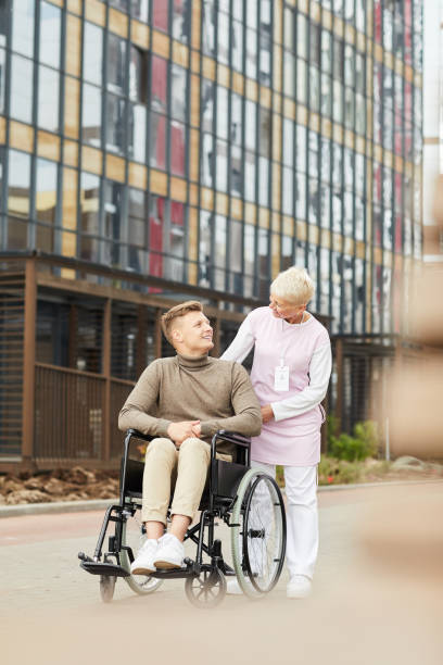 istock Positive kind mature nurse in comfortable uniform chatting with handicapped patient while they walking in yard of medical center 1168910341