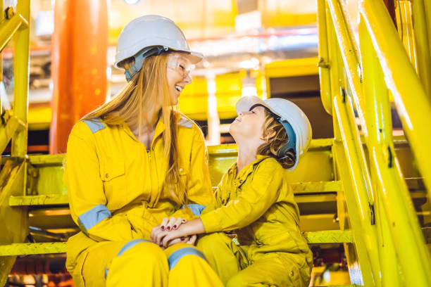 Young woman and a little boy are both in a yellow work uniform, glasses, and helmet in an industrial environment, oil Platform or liquefied gas plant stock photo