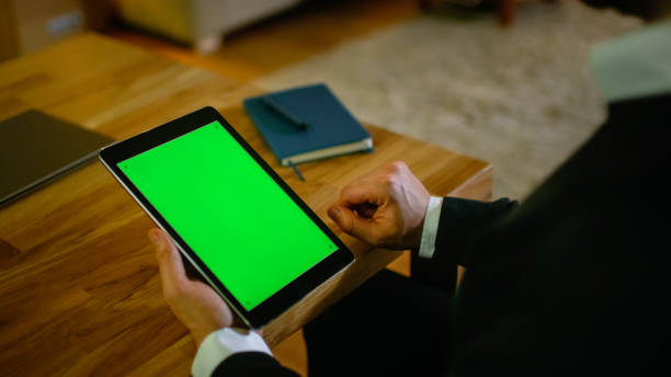 istock At Home Man Sits at His Desk and Holds Tablet Computer with Green Screen on it. His Apartment is Done in Yellow colours and is Warm. 1168937401