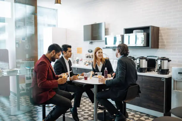 Photo of Millennial Latin coworkers taking a break at work, eating snack and chatting.