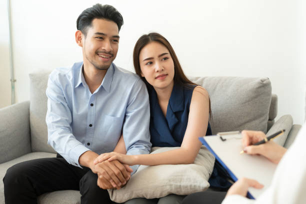 Asian couple join hand to encourage while sitting on the couch in the psychiatrist room to consult mental health problems by doctor, Health and illness concepts stock photo