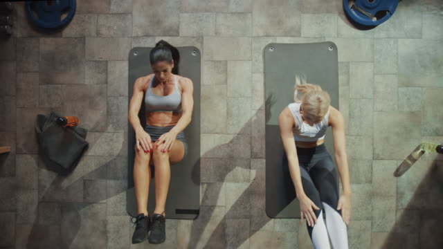 Two Professional Female Bodybuilders Doing Sit-up Exercises while Lying on the Yoga Mats in Hardcore Gym. Muscular and Athletic Beautiful Girls Workout and High Five in Celebration. Zoom Out Top View