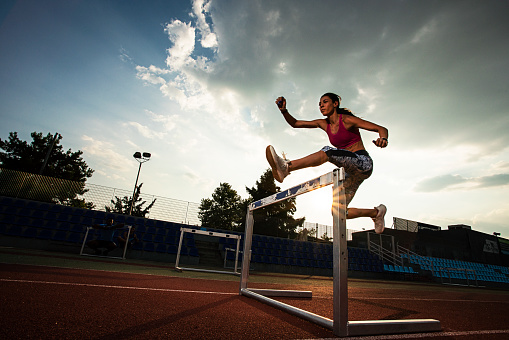 A strong woman in sports attire training for an obstacle race. She is jumping hurdles on an athletics track.