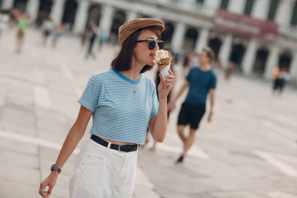 young woman eating cone gelato ice cream in hand in striped frock vest,  white pants and cap at  piazza san marco place, venice, veneto, italy - stripped shirt imagens e fotografias de stock