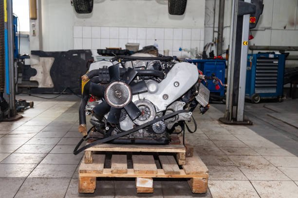 A disassembled diesel truck engine lies on wooden supports in a busy repair shop or warehouse, with a tilted truck cab visible in the background
