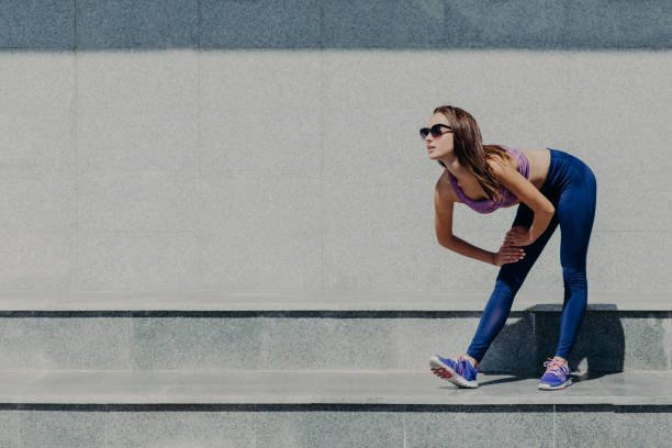 Photo of young female runner leans at knee, catches breath after jogging, dressed in top and leggings, takes break after outdoor exercise, poses outside against blank copy space for your text foto de joven corredora se apoya en la rodilla, coge el aliento después de trotar, vestido con la parte superior y leggings, toma descanso después del ejercicio al aire libre, posa fuera contra el espacio de copia en blanco para su texto
