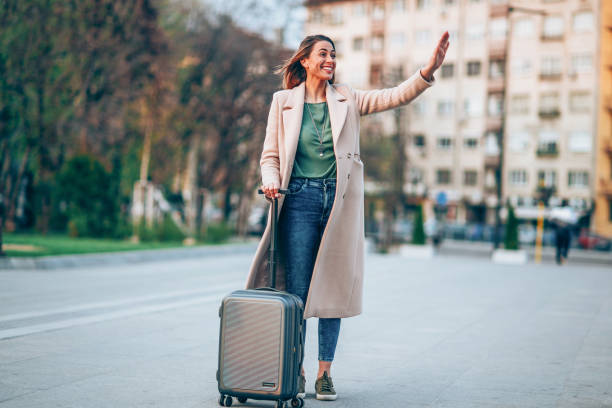 Smiling elegant woman with wheeled suitcase waving outdoors in the city