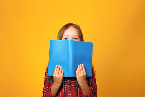 Cheerful little girl student on a yellow background. The child is hiding behind an open book and looking at the camera. Education and school concept.