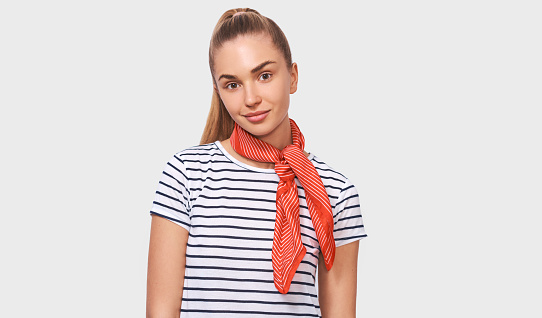 European charming young woman with ponytail hairstyle, wearing striped t-shirt, stylish red scarf on neck, looking to the camera posing over white studio background.