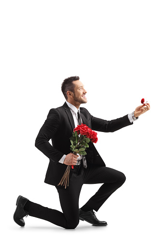 Young elegant man in a suit kneeling and holding roses and an engagement ring isolated on white background