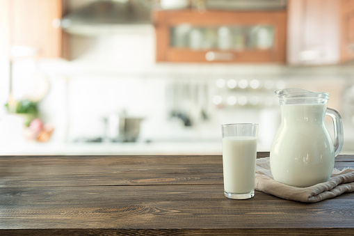 Glass of fresh milk and jug on wooden tabletop with blur kitchen as background. Space for text or display product.