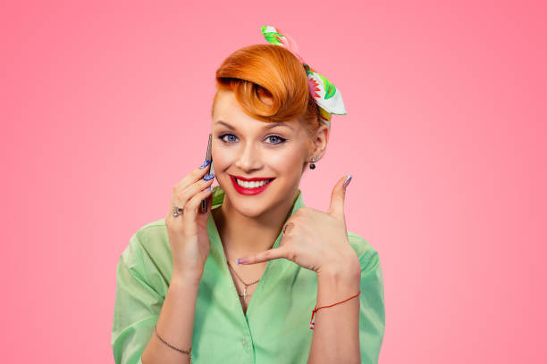 Pinup style girl talking on phone showing call me sign Call me. Closeup red head young woman pretty smiling pinup girl in green button shirt talking by phone showing call me sign hands gesture looking at you camera, retro vintage 50's hairstyle over pink pics for classic pin up poses stock pictures, royalty-free photos & images