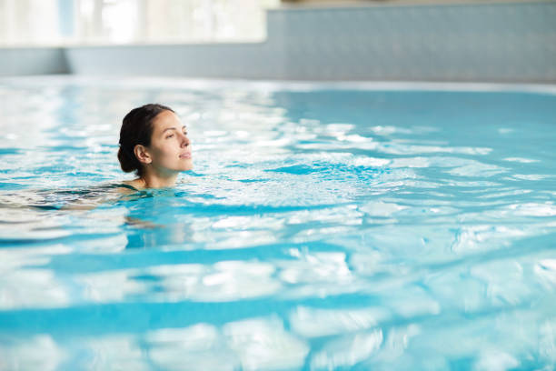 jeune femme de sourire nageant dans l'eau propre de la piscine tandis que l'attente du temps dans le club de forme physique - health spa swimming pool relaxation indoors photos et images de collection