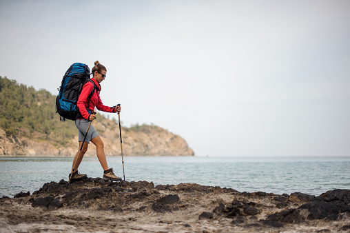 Woman in sportwear walking on the rock on the sea with hiking backpack and sticks on the beautiful background of clear sky
