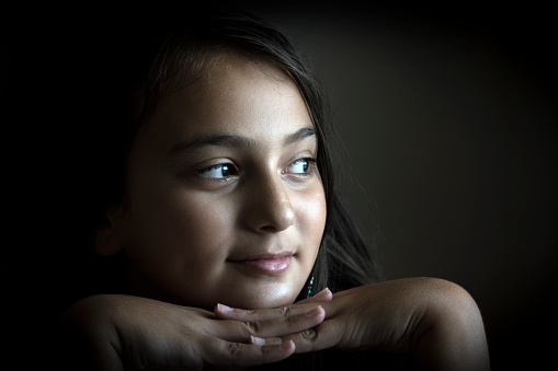 Portrait of contemplating adolescent girl having her head in her hands. She is in front of black background. The main light source is natural daylight. Shoot indoor.