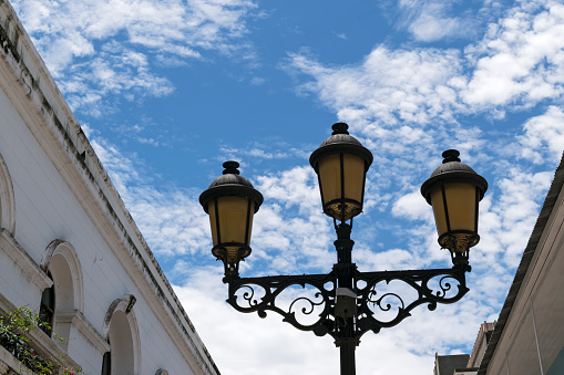 Picture of a light pole with typical lanterns in a street of the colonial zone, in the historical helmet of Santo Domingo, Dominic Republic.\nBeautiful clouds in the blue sky.