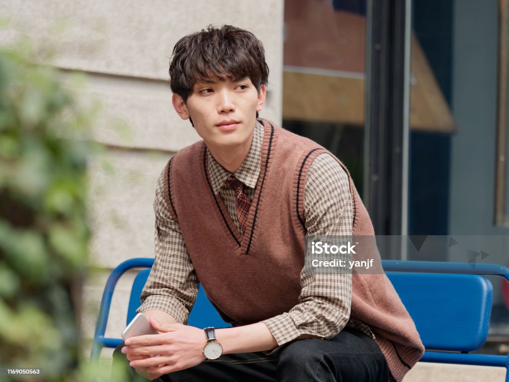 Portrait Of A Handsome Chinese Young Man With Korean Style Clothes Sitting  And Looking Away With Mobile Phone In Hand Male Fashion Cool Asian Young  Man Lifestyle Stock Photo - Download Image