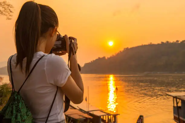 Photo of Woman tourist taking photograph with beautiful sunset view over Mekong river runs through Luang Prabang, Laos.