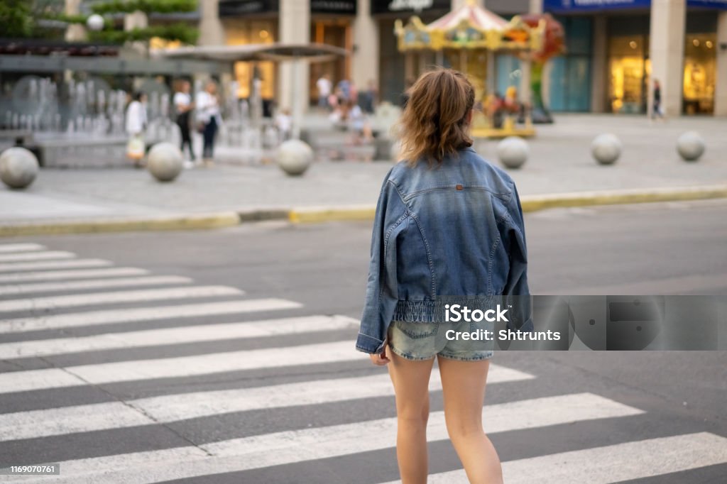 Teenage girl crosses the road. Rear view photo You can see the crosswalk, road and other details of regular urban scene. Girl dressed in a denim shorts, t-shirt and denim jacket. Place on this photo is Katerynoslavs'kyi Blvd, Dnipro city. Main colors are gray, white and blue. Photo was taken from girl's back. Rear View Stock Photo