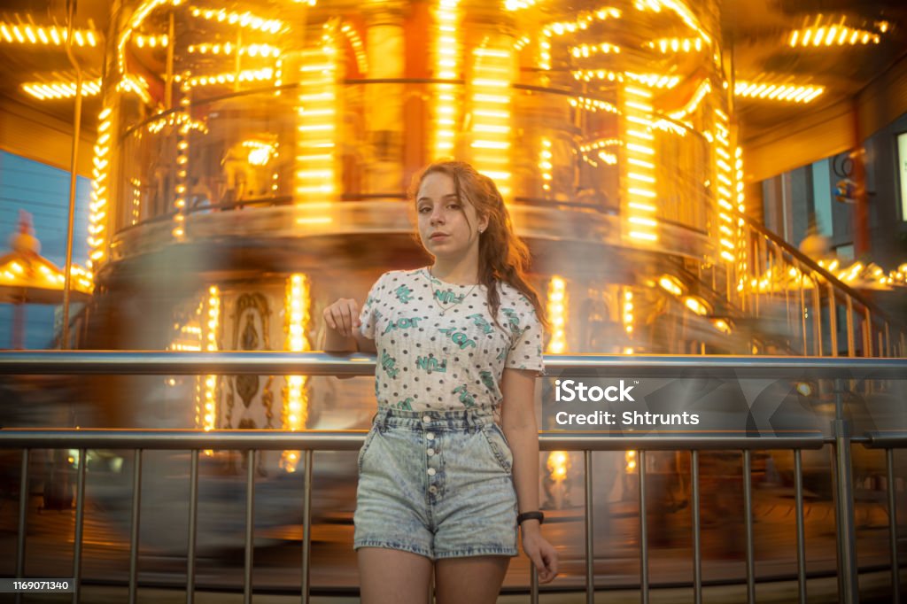 Teenage girl stands close to a spinning carousel Girl is looking down at camera. Photo was taken at evening so carousel is shining with bright orange lights. Place on this photo is central square in Dnipro city. The photo is defocused a little and contains blurred motion. It is because it was taken with long exposure and from low angle. Long Exposure Stock Photo