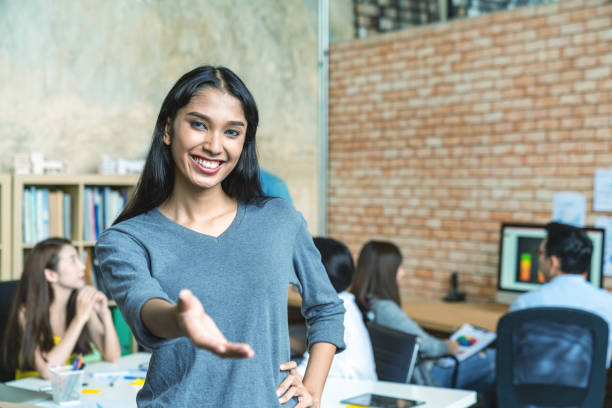 Portrait of young indian trangender extending hand and standing in creative workplace office with team work colleagues working and meeting in backgound. Welcome to my team scene and concept. Portrait of young indian trangender extending hand and standing in creative workplace office with team work colleagues working and meeting in backgound. Welcome to my team scene and concept. transgender person in office stock pictures, royalty-free photos & images