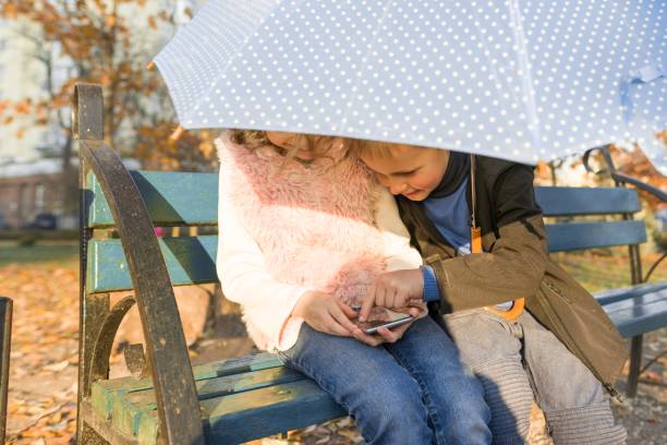 dzieci siedzące na ławce w jesiennym parku, za pomocą smartfona - couple autumn fashion male beauty zdjęcia i obrazy z banku zdjęć