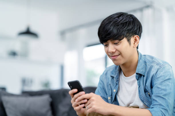 Happy asian teenager using smart phone and smiling on sofa living room at home. Asian man holding and using cellphone for searching data and social medie on internet. Teenager and Technology concept. feliz adolescente asiático usando teléfono inteligente y sonriendo en la sala de estar del sofá en casa. hombre asiático sosteniendo y usando el teléfono celular para buscar datos y medie social en internet. concepto de adolescente y tecnología.