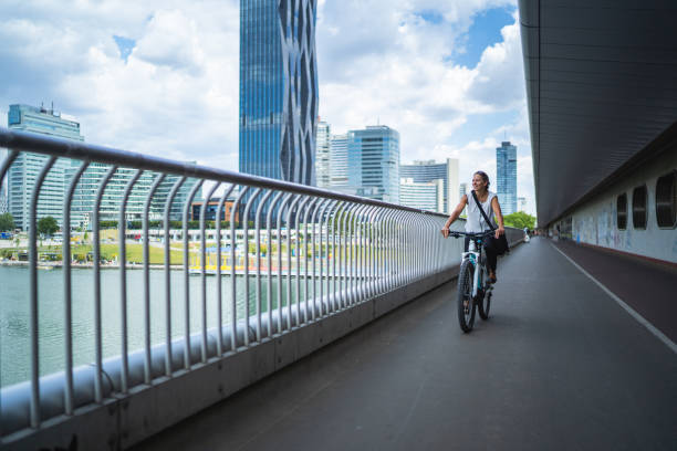 happy cycling commuter on her way to work in european city smiling 45 years old businesswoman cycling on her electric bicycle on bridge in office district in Vienna on summer day electric bicycle photos stock pictures, royalty-free photos & images