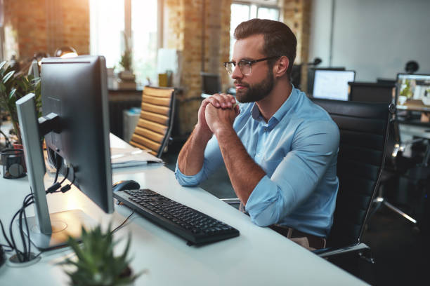 istock Thinking. Side view of young bearded man in eyeglasses and formal wear working on computer and keeping palms pressed together while sitting in the modern office 1169097231