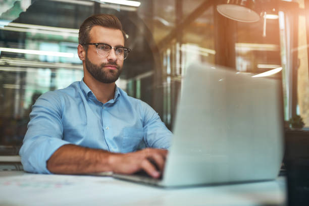 istock Comfortable working place. Young bearded businessman in eyeglasses and formal wear using laptop while sitting in modern office 1169097406