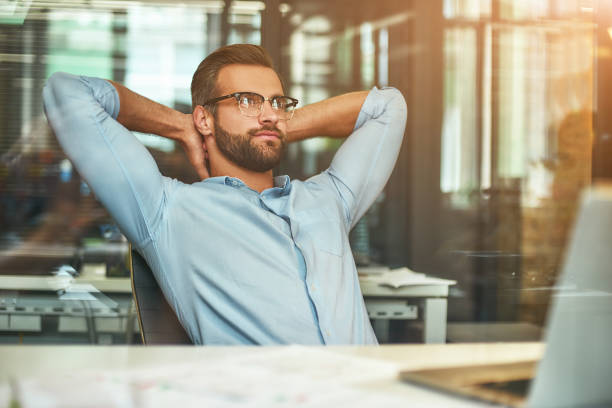 istock Relaxing. Satisfied young bearded businessman leaning back with hands behind head and thinking about something while sitting in the modern office 1169097456
