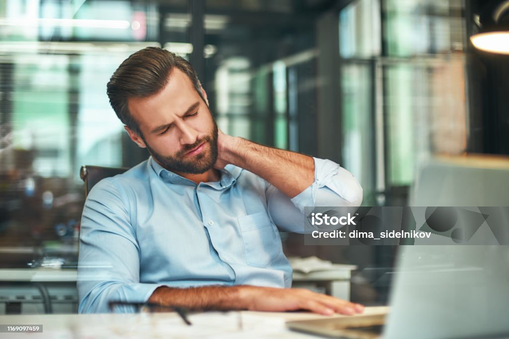 Feeling tired. Young bearded businessman in formal wear massaging his neck while sitting in the modern office Feeling tired. Young bearded businessman in formal wear massaging his neck while sitting in the modern office. Workplace. Taking a break Backache Stock Photo