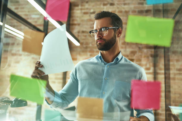 istock Creativity. Young and handsome bearded man in eyeglasses planning working process and using colorful stickers while standing in front of glass wall 1169097563