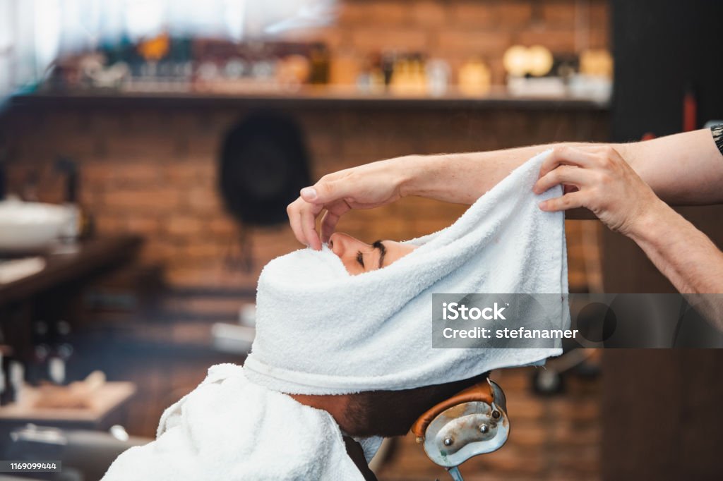 床屋は熱いタオルで男の顔を覆う。古いスタイルの理髪店で熱いと冷たい圧縮でひげを剃る伝統的な儀式。理髪店で剃る前に顔にホットタオルを持つクライアント - タオルのロイヤリティフリーストックフォト