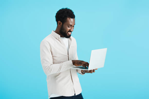 istock smiling african american man in white shirt using laptop isolated on blue 1169130188