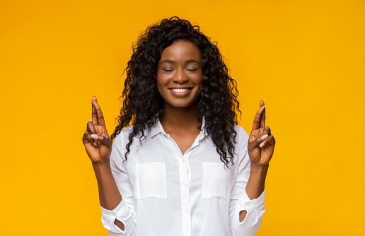 Positive Black Woman Crossing Fingers With Closed Eyes, making wish, hopes for fortune and good luck, yellow background