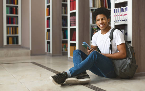 istock Cheerful afro student sitting on library floor next to bookshelves 1169135942