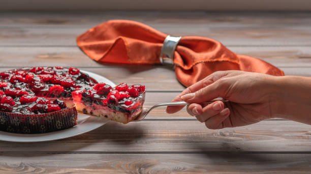 Pie with berries: raspberries, strawberries, currants, on a white plate,a woman's hand holding a piece of cake on a spatula. On a wooden background, in the background linen napkin