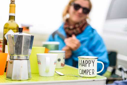 Woman camping travelling with breakfast mug and happy camper text. Woman drinking coffee or tea from special mug when camping or travelling with motor home or campervan. Vanlife camping concept.
